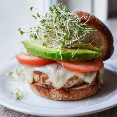 Melted Swiss cheese and creamy avocado slices top a juicy turkey burger, accompanied by crisp sprouts on a toasted bun.